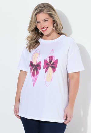 Smilende kvinde med bølget blondt hår iført en hvid T-shirt med pink spidse sko og store mørk pink sløjfe-designs.