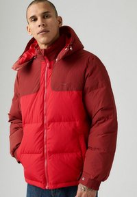 Uomo che indossa un giubbotto piumino rosso bicolor con cappuccio, in piedi con le mani nelle tasche su uno sfondo neutro.