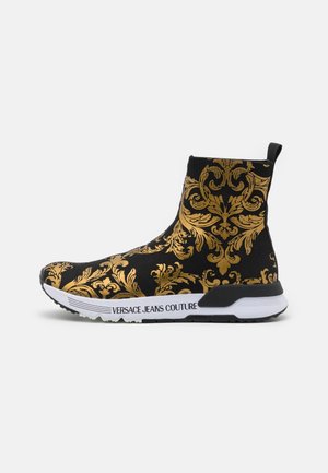 Czarny sneaker w stylu skarpetki z złotymi barokowymi wzorami kwiatowymi i białą podeszwą z napisem „Versace Jeans Couture” po boku.