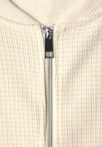 Krémově zbarvená bunda na zip s žebrovanou texturou. Má stříbrný zip a kulatý límec. Bez dalších vzorů nebo dekorací.