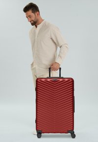 Hombre con camisa beige de pie, sonriendo, sosteniendo el asa de una gran maleta roja de carcasa dura con cuatro ruedas sobre un fondo claro.