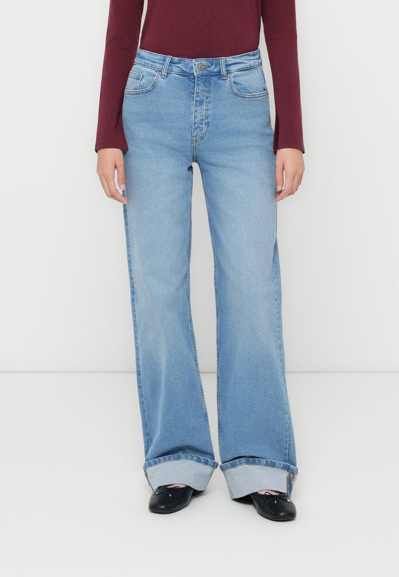 Personne portant un jean large bleu clair avec le bas retroussé, des ballerines noires et un haut bordeaux à manches longues, se tenant devant un fond blanc.