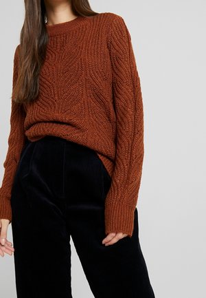 Kobieta nosząca rdzawe sweter z grubym splotem i czarne spodnie sztruksowe na jednolitym jasnoszarym tle.