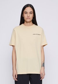 Lysegul bomulls-T-skjorte med korte ermer, rund hals og svart logo tekst på venstre brystområde. Myk tekstur, avslappet passform.