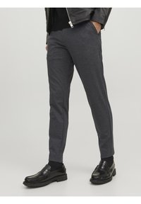 Muž oblečený v tmavě šedých slim fit kalhotách, černých ponožkách, lesklých černých mokasínech a černé kožené bundě, s rukama v kapsách.