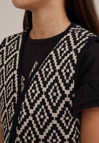 Czarna i beżowa kamizelka w wzór diamentów, wykonana z teksturowanej dzianiny, nałożona na czarną koszulkę z krótkim rękawem.