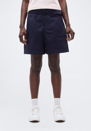 Personne portant un short bleu marine avec ceinture, des chaussettes blanches montantes et des baskets blanches, debout devant un fond clair uni.