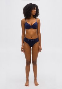 Γυναίκα όρθια, ξυπόλυτη με φόντο ένα απλό σκηνικό, φοράει μπλε σουτιέν και εσώρουχο με το λογότυπο "POLO" στη ζώνη, κοιτάζοντας προς τα αριστερά της.