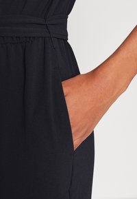 Prenda de tela negra con un acabado texturizado, que cuenta con un bolsillo lateral y un lazo de tela en la cintura para un ajuste regulable.