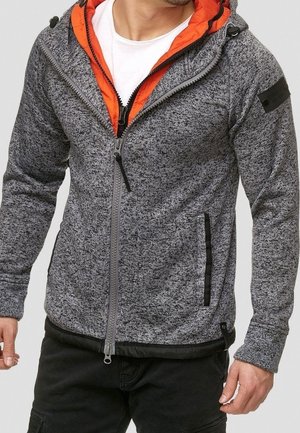 Homme portant un sweat à capuche gris zippé avec une doublure orange sur une chemise blanche et un pantalon noir, debout, les bras détendus.