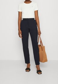 Bílá bavlněná tričko, námořnické kalhoty na míru, černé sandály na pásky a hnědá semišová taška. Má vodorovné pruhy na pase.