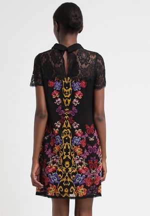 Kobieta z włosami upiętymi w kok, ubrana w czarną sukienkę z koronkowymi rękawami i kolorowym haftowanym wzorem kwiatów oraz motyli na plecach.