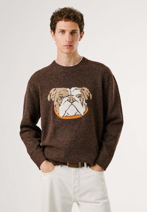 Braun gestrickter Pullover mit einem Bulldoggen-Motiv in Creme und Orange. Runder Hals und gerippte Bündchen. Getragen mit hellen Hosen.