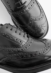 Čierne kožené brogue topánky s perforovanými detailmi, vystúpenými stehmi a textúrovanou podrážkou. Majú klasický šnurovací dizajn.