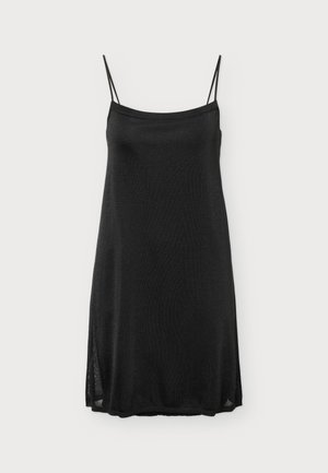 Femme portant une mini-robe noire sans manches avec de fines bretelles et des bottes en cuir noires montant jusqu'aux genoux, debout devant un fond uni.
