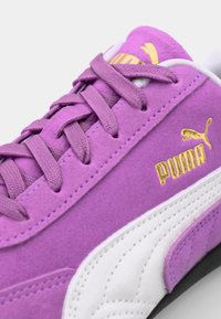 Violet samettikengät, joissa on valkoisia nahkayksityiskohtia, pyöristetty kärki, litteät nauhat ja kultainen logopainatus. Teksturoitu pinta ja ompelukuviot.