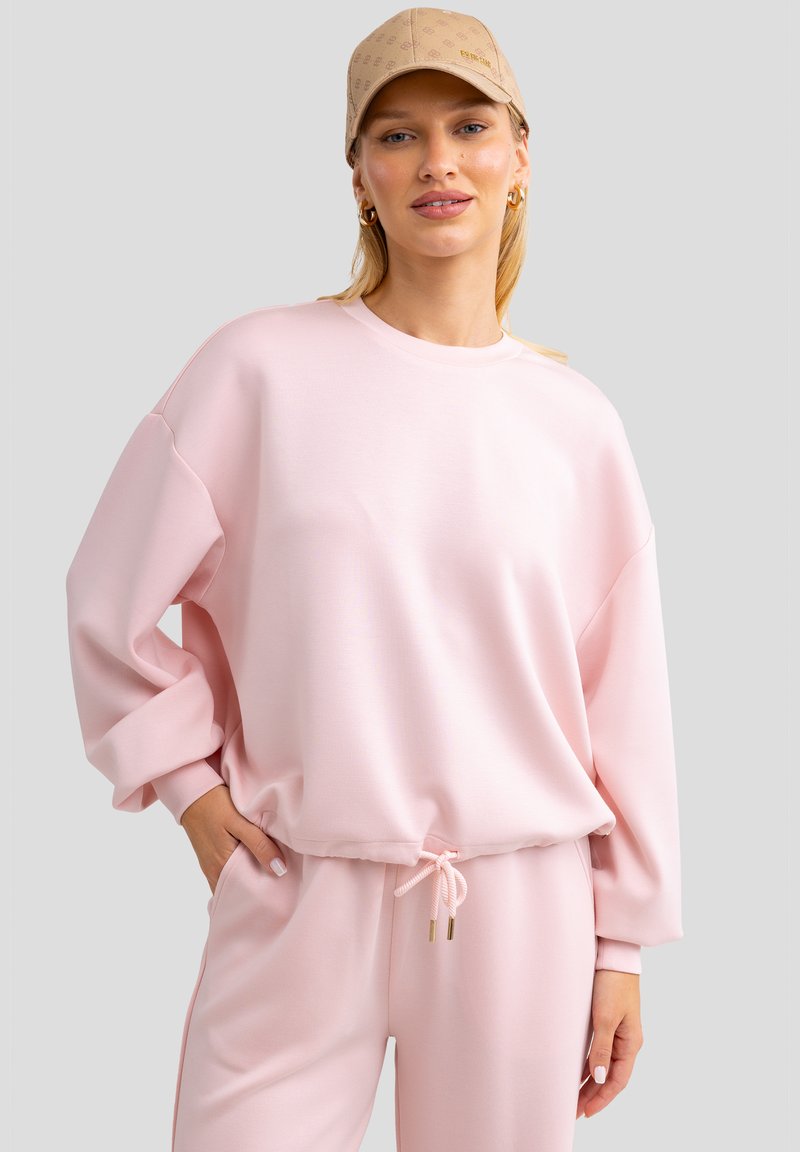 Sudadera rosa claro de corte relajado, con costuras de hombro caídas y un dobladillo con cordón. Combina con pantalones a juego y una gorra color arena.
