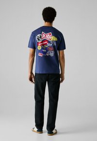 Navy blauwe katoenen t-shirt met een grote grafiek van logo's en patches op de achterkant. Zwarte jeans en meerkleurige schoenen maken de outfit compleet.