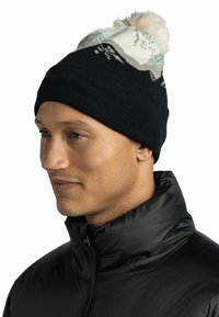 Gorro de punto negro con un patrón de montañas en blanco y azul, rematado con un pompón esponjoso. El modelo lleva una chaqueta negra con cremallera.