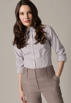 Kobieta z długimi falowanymi ciemnymi włosami, ubrana w pasiastą koszulę na guziki i beżowe spodnie z wysokim stanem, stojąca z jedną ręką w kieszeni.