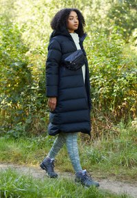 Dlhý čierny prešívaný kabát s kapucňou, skombinovaný s mentolovo zelenými vzorovanými legínami a šedými turistickými topánkami. Čierna crossbody taška nosená na páse.