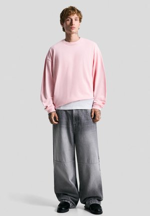 Lys lyserød oversized sweatshirt med rund halsudskæring, båret over en hvid skjorte. Sammen med brede, falmede grå denim bukser og sorte sko.