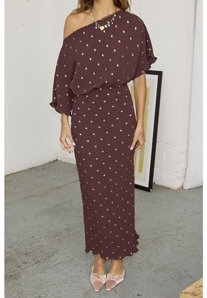 Kobieta ubrana w bordo sukienkę na jedno ramię z złotymi grochami i różowymi butami w szpic, stojąca wewnątrz przy oprawionym dziele sztuki.