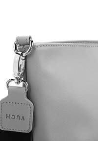 Nahaufnahme einer grauen Lederhandtasche, an deren Ecke die genähten Kanten und ein Metallclip zu sehen sind, der einen Riemen mit einem kleinen rechteckigen Anhänger verbindet, auf dem "VUCH" eingeprägt ist.