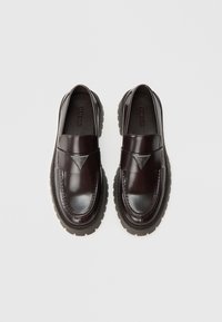 Mocasini din piele lustruită burgundy, cu talpă groasă din cauciuc negru, având un logo triunghiular și cusături decorative.