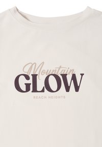Bílý bavlněný tričko s výrazným grafickým textem v tmavě hnědé a světlé béžové barvě: "Mountain GLOW" a "REACH HEIGHTS" pod ním v moderním písmu.
