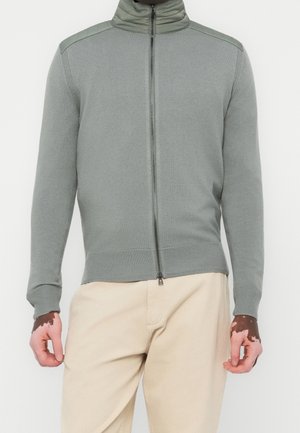 Mężczyzna w jasnoszarej bluzie na zamek z wysokim kołnierzem i beżowych spodniach, stojący na jednolitym białym tle.