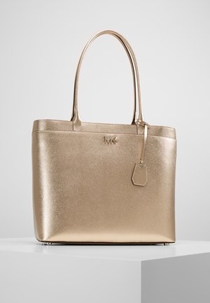 Sac fourre-tout en cuir métallisé doré avec poignées doubles, logo MK à l'avant, et une étiquette en cuir suspendue, posé sur une surface blanche.