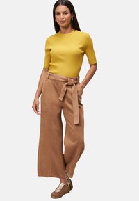 Geel geribbelde top met korte mouwen, gecombineerd met hooggeknoopte, bruin brede broek met een strikceintuur. Schoeisel is bruin sneakers met accentdetails.