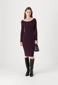 Long-sleeve, fitted dress in deep purple fabric, featuring an off-shoulder neckline and subtle ruching. Paired with black knee-high boots.