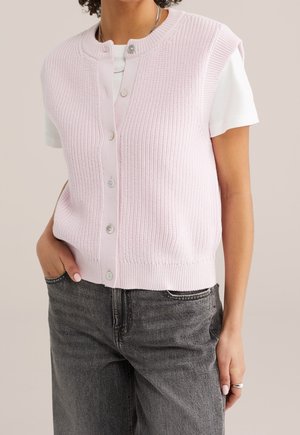 Modèle portant un gilet en maille côtelée rose pâle, boutonné devant, sur un T-shirt blanc et un jean gris foncé.
