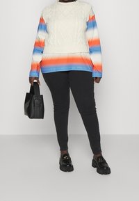 Jersey de punto con diseño de cable texturizado en color crema, con rayas degradadas en azul y naranja. Combinado con pantalones negros ajustados y zapatos gruesos.