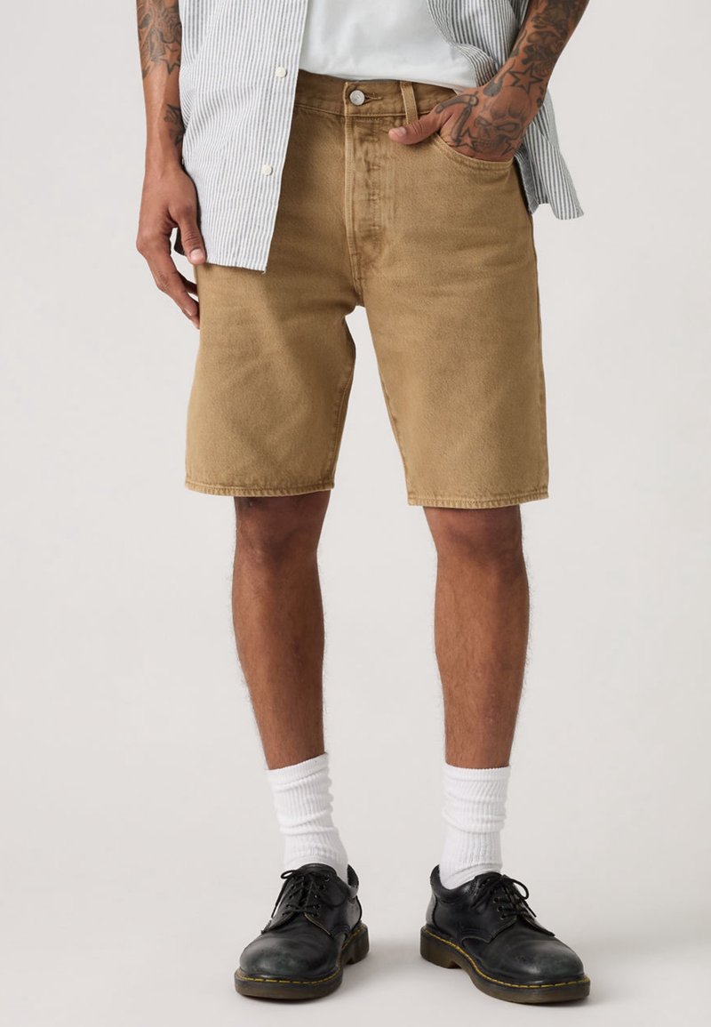 Homme portant un short beige arrivant aux genoux, des chaussettes blanches, des chaussures noires et une chemise rayée partiellement déboutonnée avec une main dans la poche.