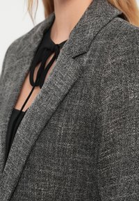 Pilkas švarkas iš tekstūruotos medžiagos, su įpjauta apykakle ir matomomis siūlėmis. Po juo - juoda palaidinė su kaklaraiščio detalė.