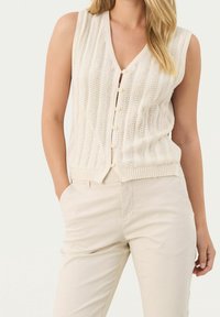 Ivoren gebreide mouwloze cardigan met verticale patronen en een V-halslijn. Voorzien van knoopsluitingen en een geribbelde zoom. Gecombineerd met lichtbeige broeken.