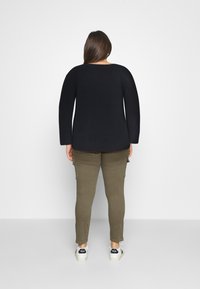 Femme aux longs cheveux bruns portant un pull noir, un pantalon cargo vert olive et des baskets blanches, debout face à un mur blanc.
