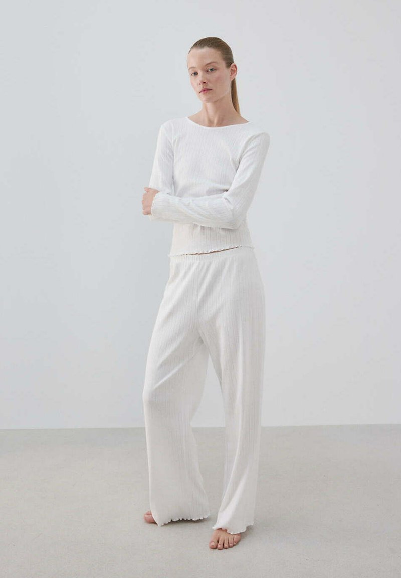 Femme aux cheveux attachés debout pieds nus, portant un haut blanc à manches longues et un pantalon blanc à jambes larges sur un fond clair uni.