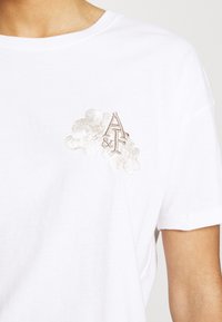 Hvid bomulds t-shirt med et diskret broderet design med "A&F" og blomsterdetaljer i lys tan. Klassisk rund halsudskæring og afslappet pasform.