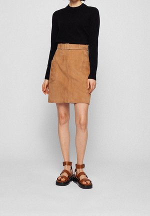 Femme portant un haut noir à manches longues, une mini-jupe en daim beige ceinturée avec poches, et des sandales en cuir marron à boucle se tenant devant un fond uni.