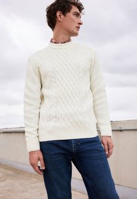 Weißer Kabelstrickpullover mit strukturierten Mustern und geripptem Saum, kombiniert mit dunkelblauen Jeans, vor einem bewölkten Hintergrund präsentiert.