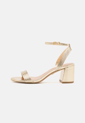 Guldmetallic åben sandal med blokhæl, ankelrem med guldlås og parallelle remme detaljer på tå og hæl.