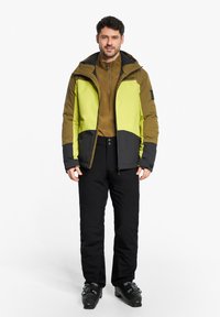 Men's insulated jacket in lime green, brown, and black, with a front zipper, hood, and multiple sections. Paired with black pants and snow boots.