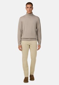 Beige turtleneck džemperis ar ribotu apkakli un apakšu, apvienots ar gaiši beigu biksēm un brūnām zamša kurpēm, stāvot uz vienkrāsaina fona.