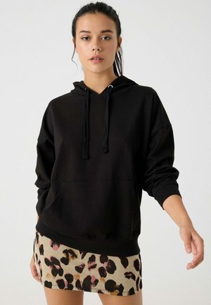 Sudadera negra con bolsillo canguro y cordones ajustables; combinada con una falda corta beige que presenta un patrón de estampado de leopardo multicolor.