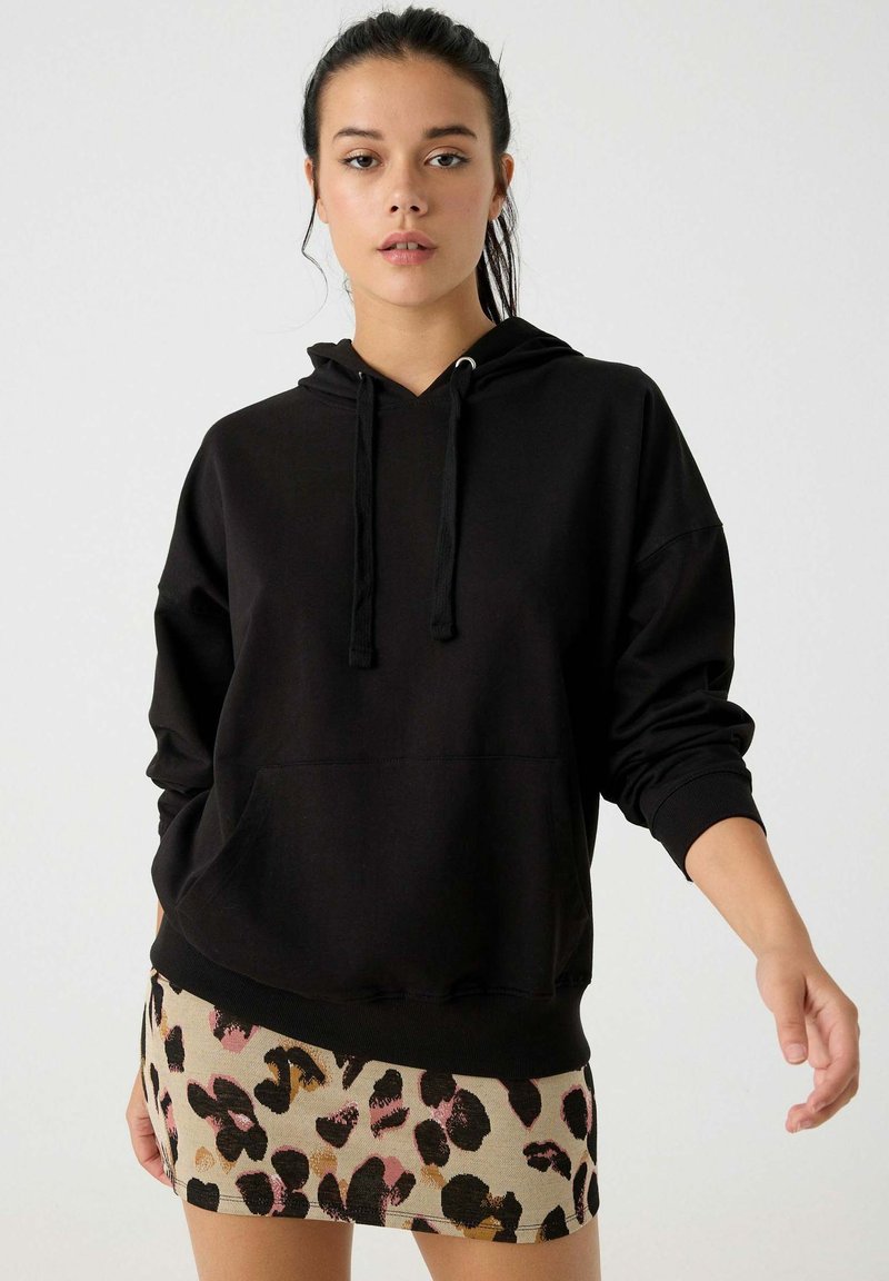 Sudadera negra con bolsillo canguro y cordones ajustables; combinada con una falda corta beige que presenta un patrón de estampado de leopardo multicolor.
