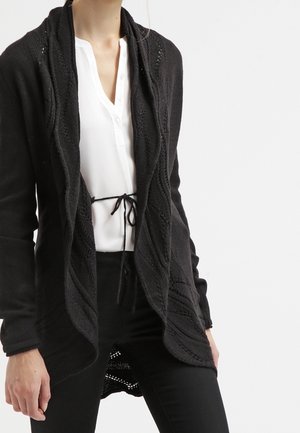 Zwarte gebreide cardigan met een open voorkant, voorzien van een gestructureerd patroon en een sjawl-kraag. Gecombineerd met een witte blouse en zwarte broek.